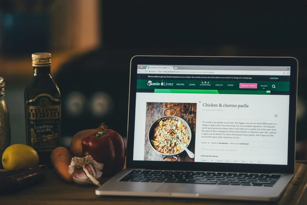 An open laptop rests on a wooden counter displaying a Jamie Oliver recipe page for chicken and chorizo paella. Beside it sit fresh cooking ingredients, including a red bell pepper, garlic, a lemon, and a bottle of Filippo Berio olive oil.