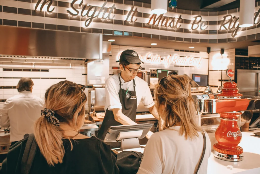 A young male employee wearing glasses and an apron assists two female customers at a diner counter. Overhead sign text reads "In Sight It Must Be Right" above the stainless-steel kitchen station.