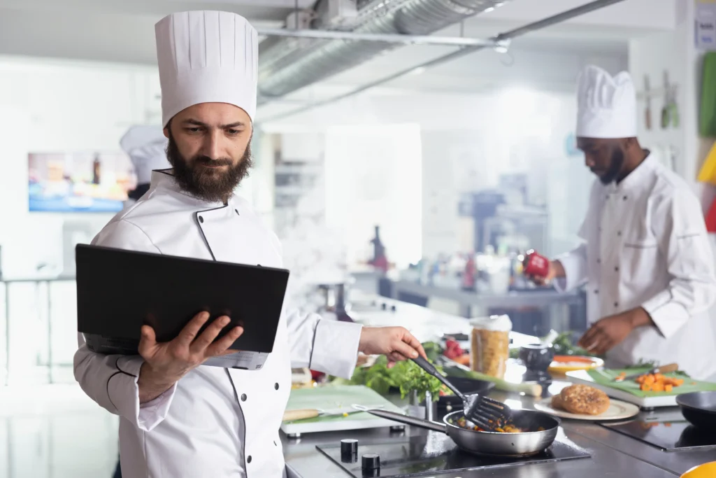 A bearded male chef wearing a white uniform and toque holds a laptop while stirring food in a pan. In the background, another chef in matching attire works at a busy restaurant kitchen counter.