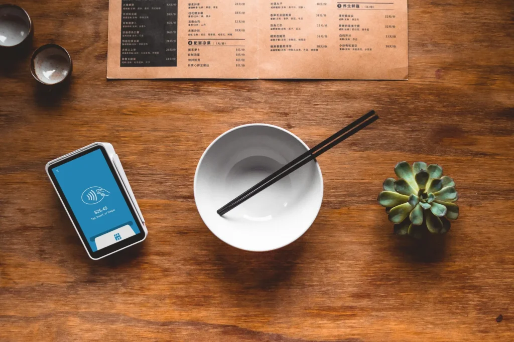 A flat-lay view of a wooden dining table features a payment terminal displaying a contact-less prompt on the left and an empty white bowl with black chopsticks rested across it in the center. At the top sits an open restaurant menu printed in Chinese, flanked by two small teacups on the left and a small succulent plant on the right.