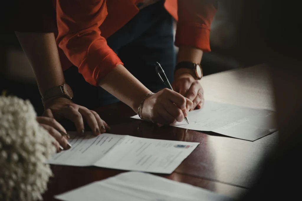 A person wearing an orange blazer leans over a wooden table to sign a paper document with a pen. Another document rests nearby alongside a cluster of light-colored flowers in a warmly lit, moody setting.