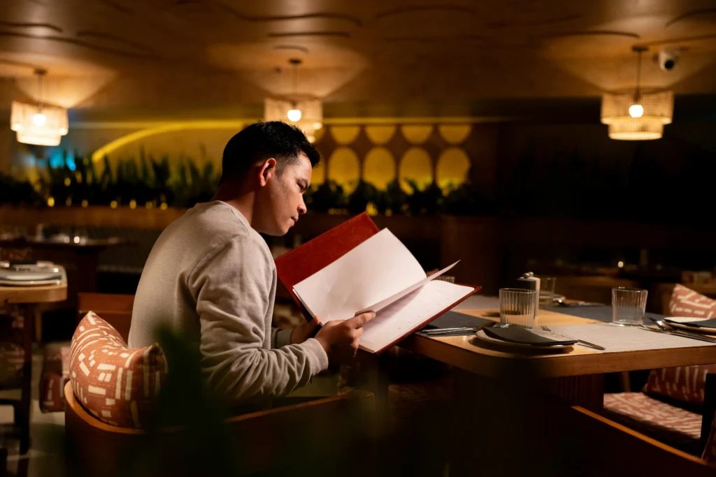 A man sits alone at a dining table in a warmly lit, upscale restaurant while looking intently at an open menu. The ambient lighting overhead casts a cozy, intimate glow across the sophisticated interior space.