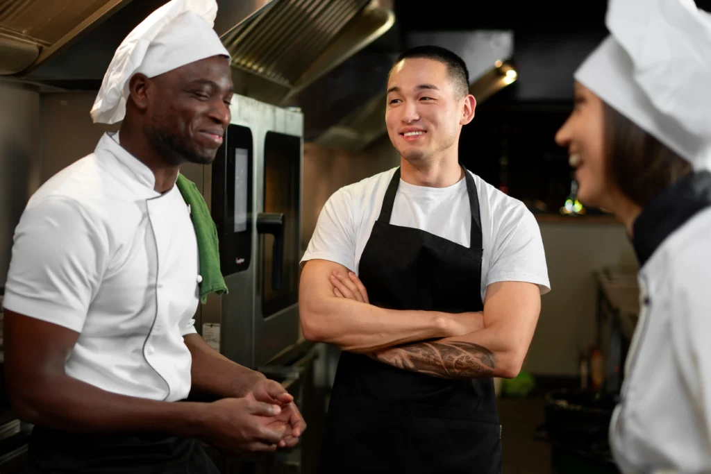 Three diverse kitchen staff members wearing chef uniforms stand together and smile while conversing in a commercial kitchen. One chef stands with his arms crossed in the center while sharing a lighthearted moment with his colleagues.