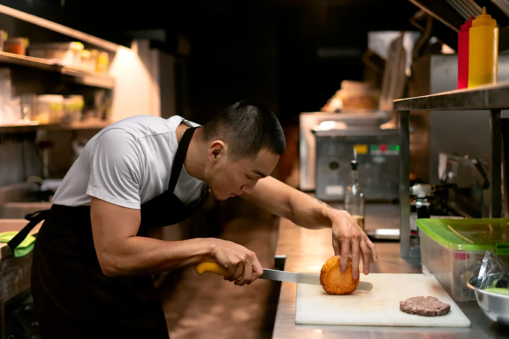 A male chef wearing a white t-shirt and black apron carefully slices a burger bun on a cutting board using a serrated knife. He leans intently over the stainless steel kitchen counter with a raw meat patty resting nearby.