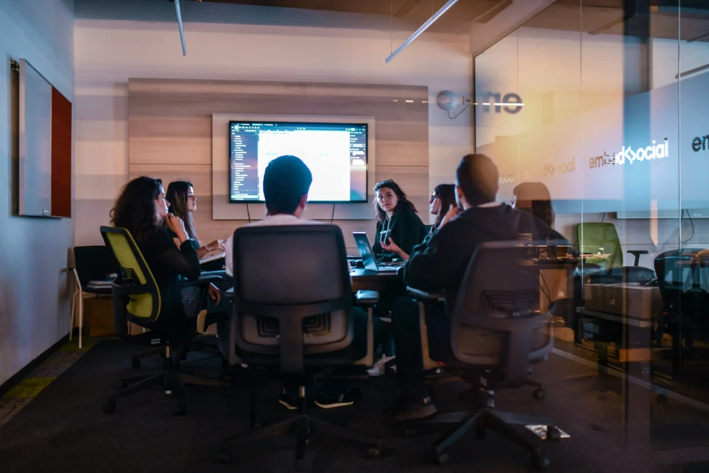A group of colleagues sits around a conference table engaged in a meeting inside a modern office room. A large screen displaying software code and data is mounted on the wall in front of them.
