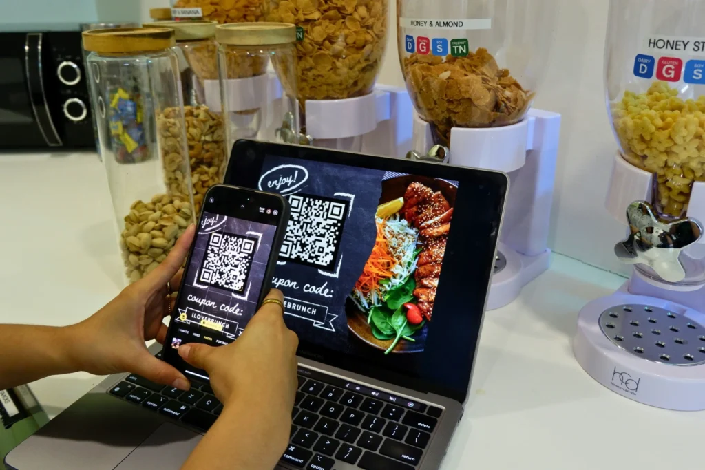 A person holds a smartphone over an open laptop screen to scan a QR code for a food discount coupon code. In the background, clear countertop cereal dispensers and snack jars are arranged near the workspace.
