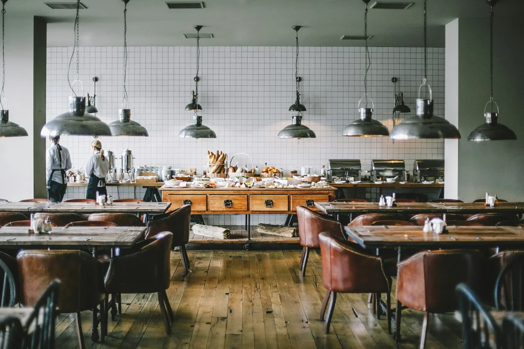 A spacious, rustic-industrial restaurant dining room features wooden tables, leather armchairs, and metal pendant lights hanging from above. In the background, staff stand near a breakfast buffet spread positioned against a tiled white wall.