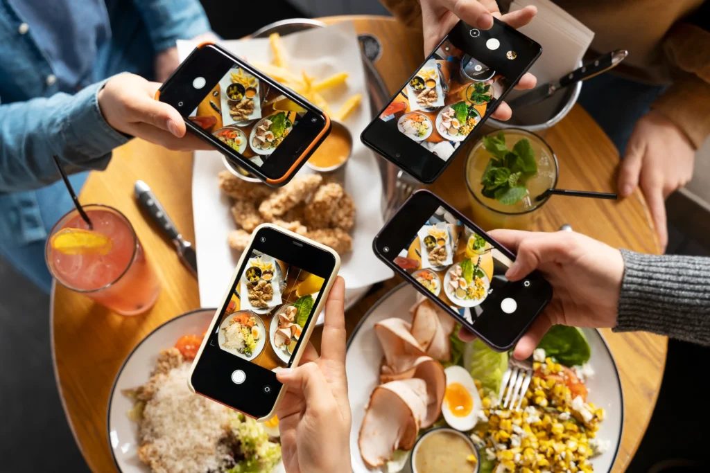 An overhead view shows four people holding their smartphones to take pictures of a shared table filled with food and drinks. The wooden table is spread with colorful dishes, including fried chicken, salads, fries, and iced cocktails.