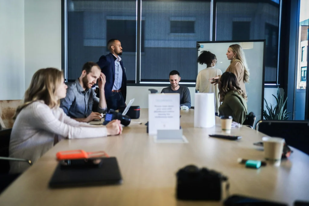 A diverse group of colleagues gathers around a large wooden table inside a bright, modern office conference room. Several people collaborate on laptops and engage in discussions while others converse near a whiteboard in the background.