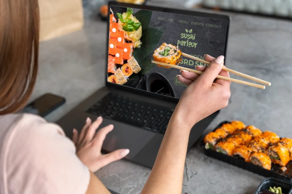 A woman sits at a table holding a piece of sushi with chopsticks in front of an open laptop showing a restaurant website called "Sushi Parlour." A takeout container filled with sushi rolls sits nearby on the counter.