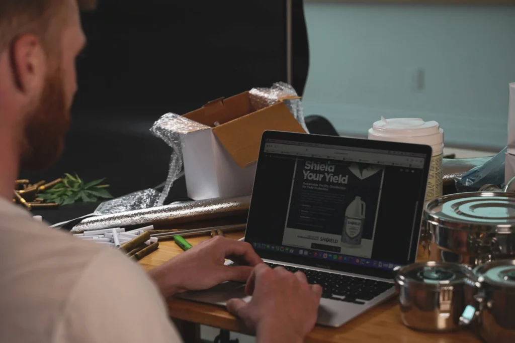 A man works on an open laptop displaying a "Shield Your Yield" webpage. His cluttered desk is filled with packaging materials, metal containers, and various supplies.