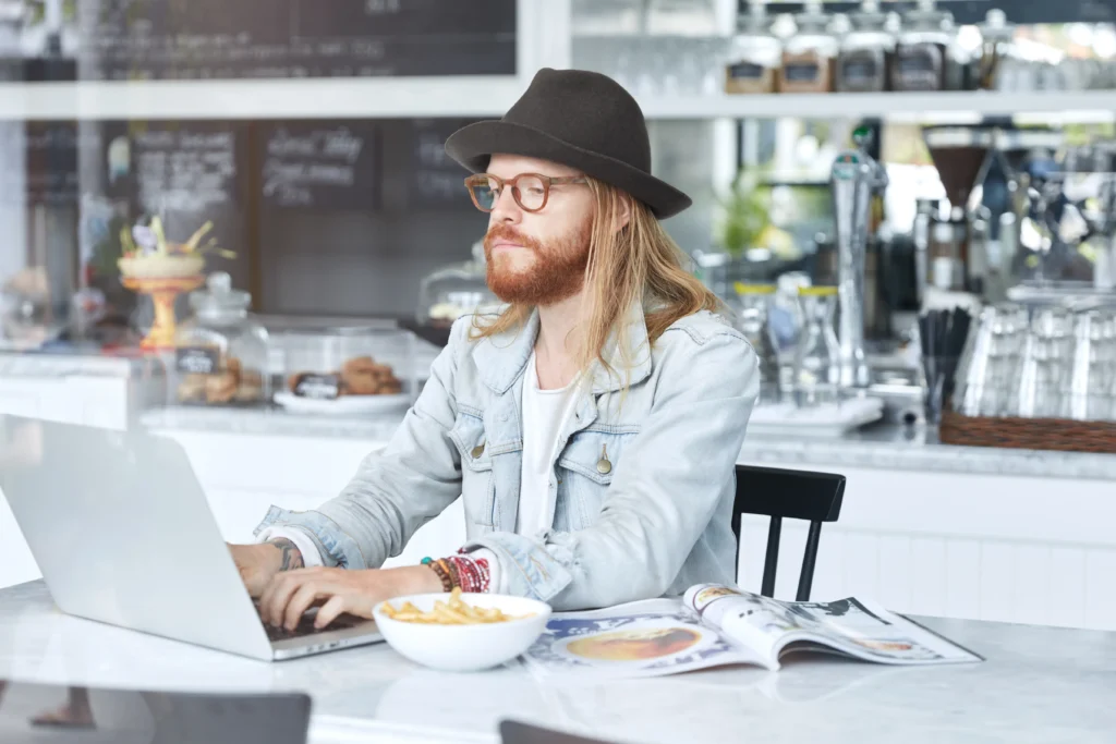 A stylish man with long blonde hair, a red beard, and a brown fedora hat works intently on a laptop at a café counter. Next to his computer sits a bowl of French fries and an open magazine.