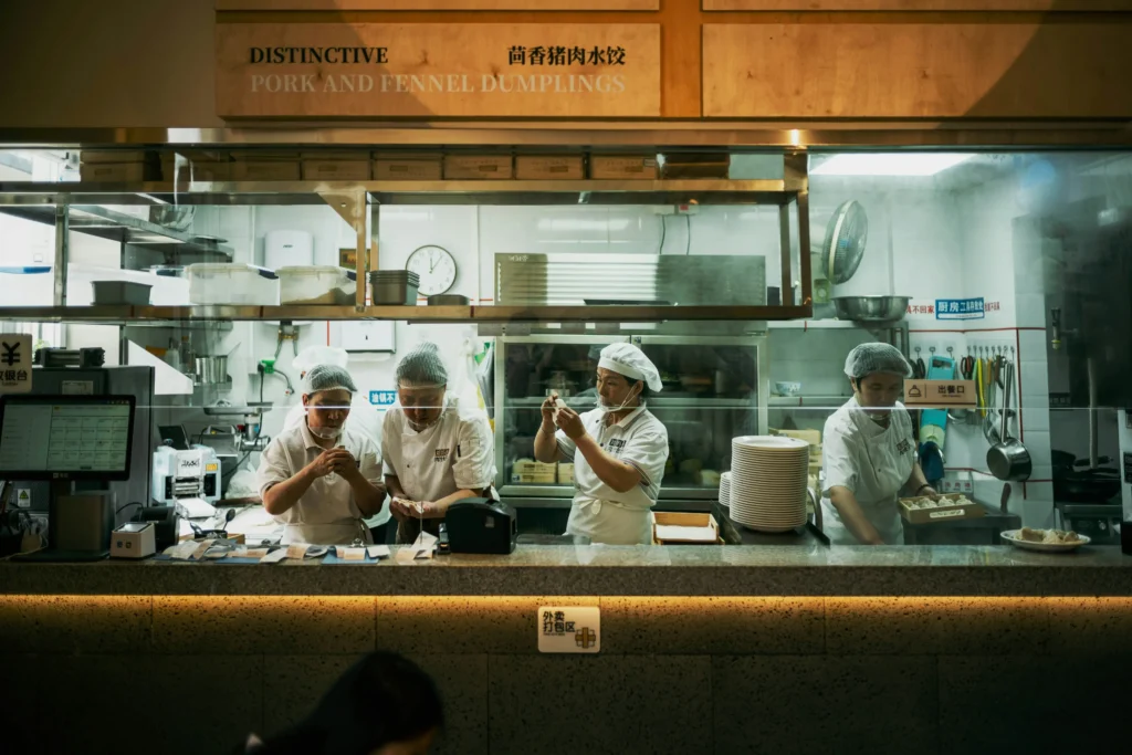 Four kitchen staff wearing white uniforms and hairnets work behind an open restaurant counter. A sign above them advertises "Distinctive Pork and Fennel Dumplings" in English and Chinese.