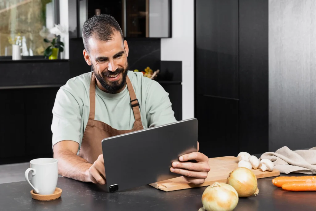 A smiling man wearing a brown apron holds a tablet computer in a modern kitchen. Fresh vegetables and a white coffee mug sit on the counter in front of him.