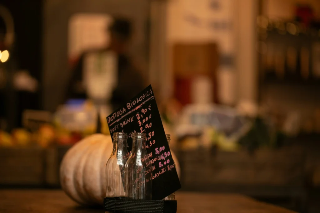A small handwritten chalkboard menu rests inside glass bottles on a wooden counter next to a large pumpkin. The warm, dimly lit cafe background creates a cozy, rustic atmosphere.