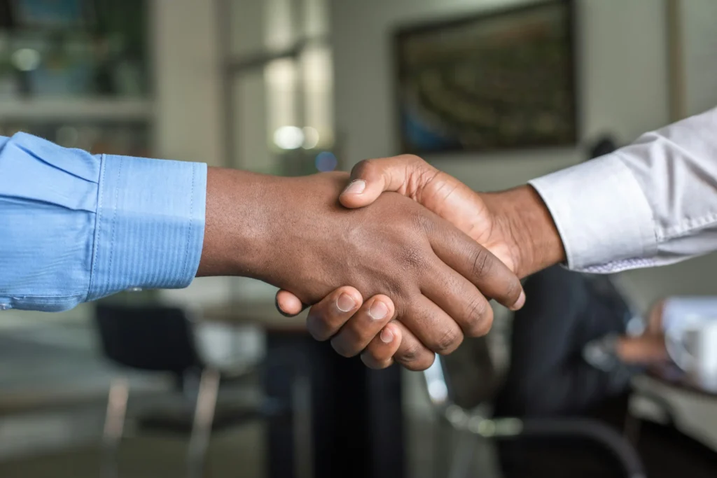A close-up shot of two people shaking hands in a professional office setting. One person wears a blue button-down shirt while the other wears a white long-sleeved shirt.