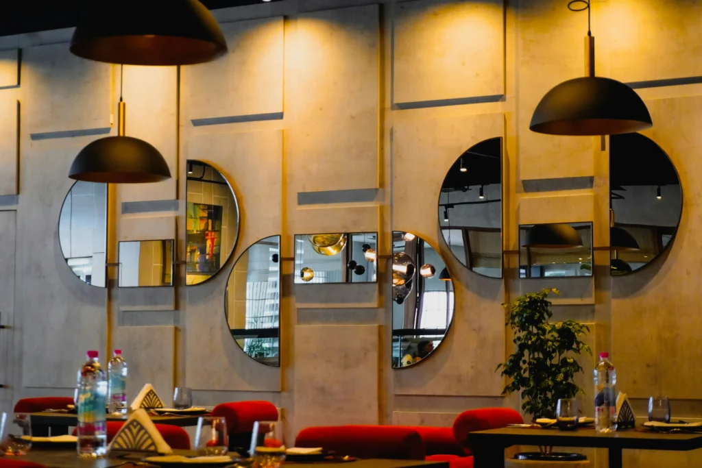 A modern restaurant interior featuring paneled walls mounted with stylish semicircular and rectangular mirrors. Warm black dome pendant lights hang above dining tables set with red seats and glassware.