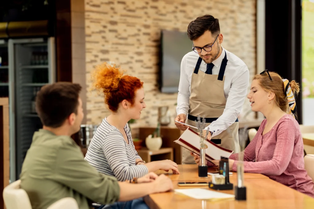 A young male waiter wearing glasses and an apron stands at a restaurant table, presenting and pointing to a menu for a seated group of friends. The three guests look attentively at the menu as they decide on their orders in a brightly lit cafe setting.