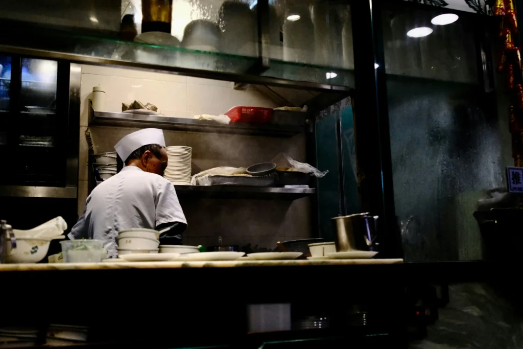 A chef wearing a white paper hat and uniform works in a dimly lit, narrow kitchen stall. Steam rises near stacks of plates and food preparation supplies arranged on shelves above the counter.