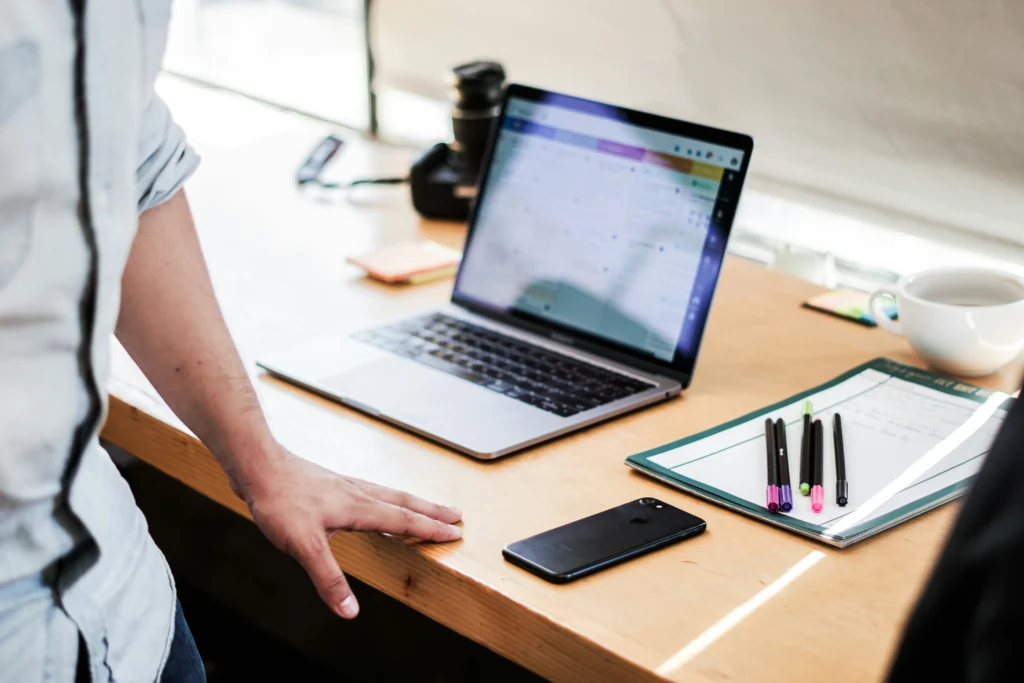A person stands next to a wooden desk featuring an open laptop displaying a colorful digital interface. Surrounding the laptop are various work items, including a smartphone, a clipboard with colorful pens, sticky notes, and a coffee cup.