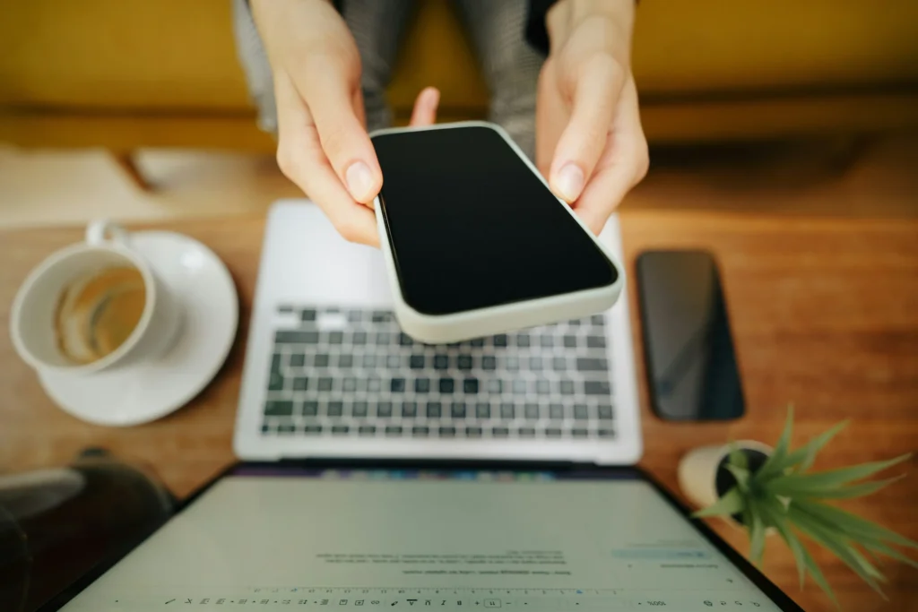 A top-down view shows a person holding out a smartphone with a blank black screen toward the camera. In the background, an open laptop, a cup of coffee, and a small potted plant sit on a wooden table.