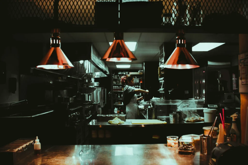 A chef stands at a food preparation station inside a dimly lit restaurant kitchen. Three copper cone heat lamps hang directly above the stainless steel counter, casting a warm glow over the workspace.