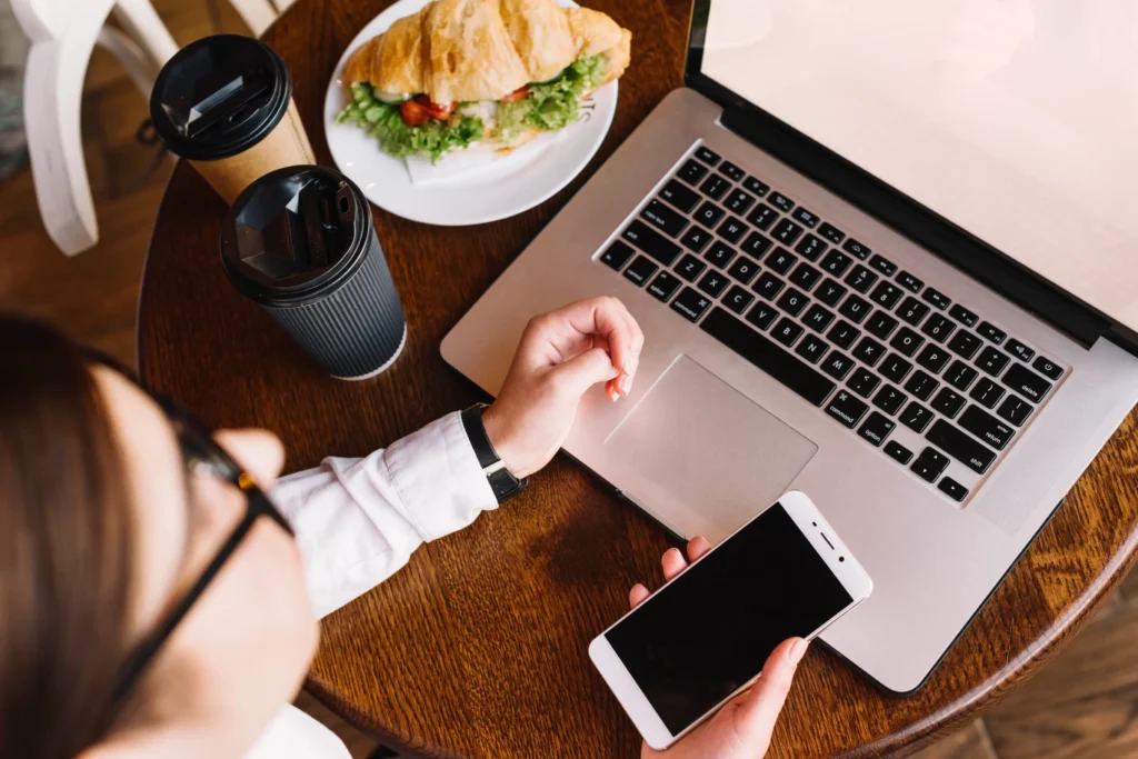 Over-the-shoulder view of a woman working at a wooden cafe table with an open laptop and holding a smartphone. Next to her devices sit two coffee cups and a plate with a croissant sandwich.