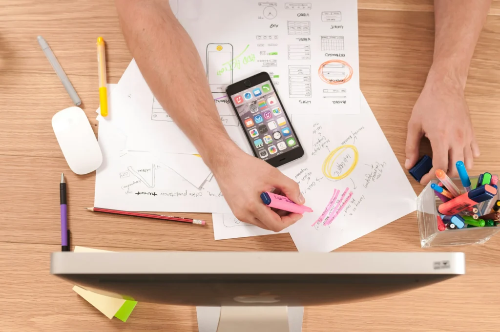 An overhead view shows a designer working at a wooden desk filled with app wireframe sketches, colorful highlighters, and a smartphone. They use a pink highlighter on a document while sitting behind a computer monitor.