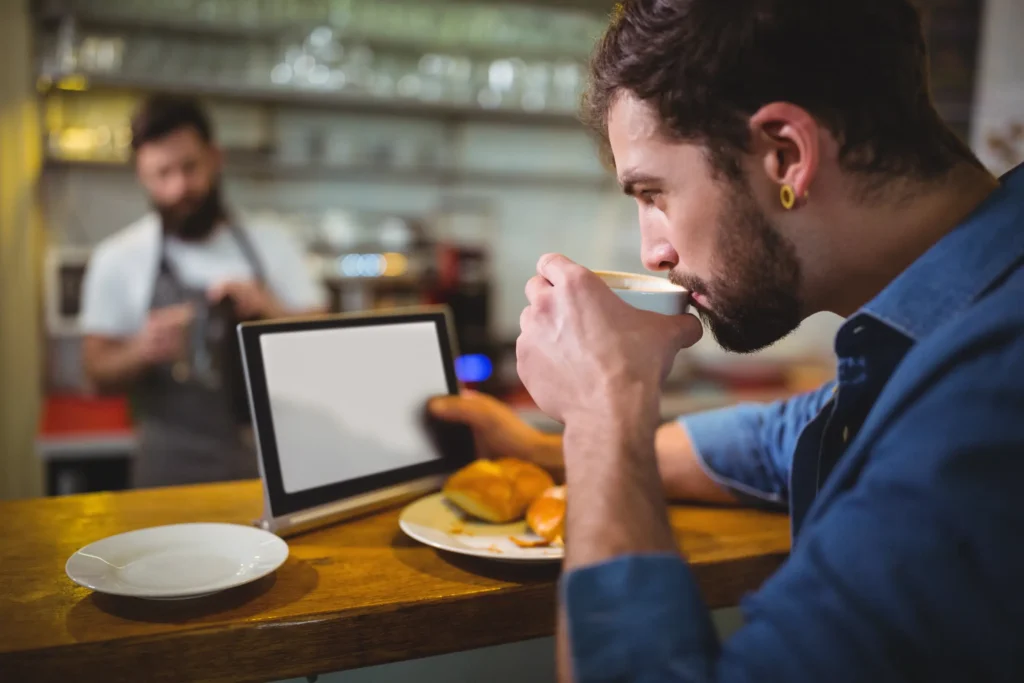 A bearded man takes a sip from a coffee cup while looking at a blank digital tablet on a wooden cafe counter. In the blurred background, a barista works behind the bar.
