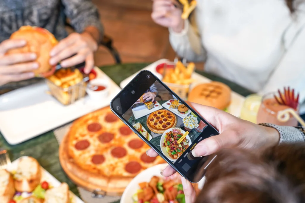 A person holds a smartphone over a dining table, taking a photo of a meal spread featuring pepperoni pizza, burgers, fries, and salads. Around the table, friends enjoy their food during a social gathering.