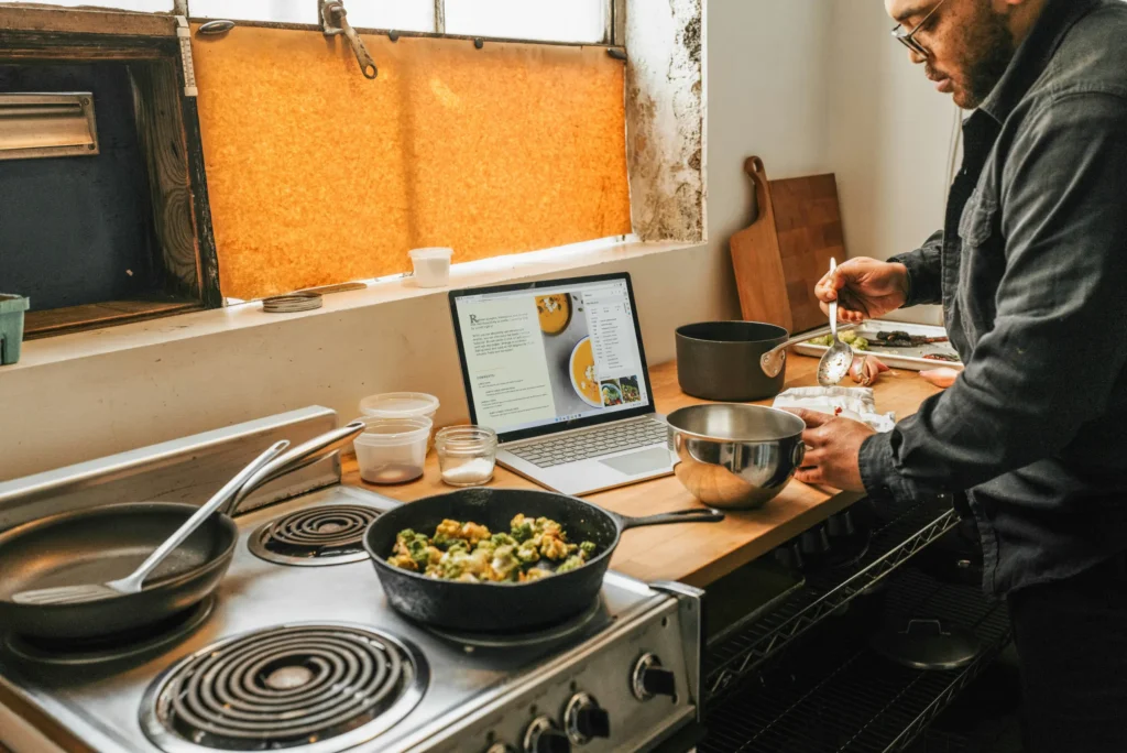 A man wearing glasses stands at a kitchen counter, using a spoon to prepare food with a recipe open on a nearby laptop. Skillets rest on the adjacent stovetop alongside various cooking ingredients and utensils.
