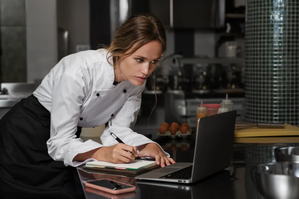 A female chef in a white chef coat leans over a kitchen counter while focused on her open laptop. She holds a pen to take notes in a notebook, with ingredients like fresh eggs visible in the background.