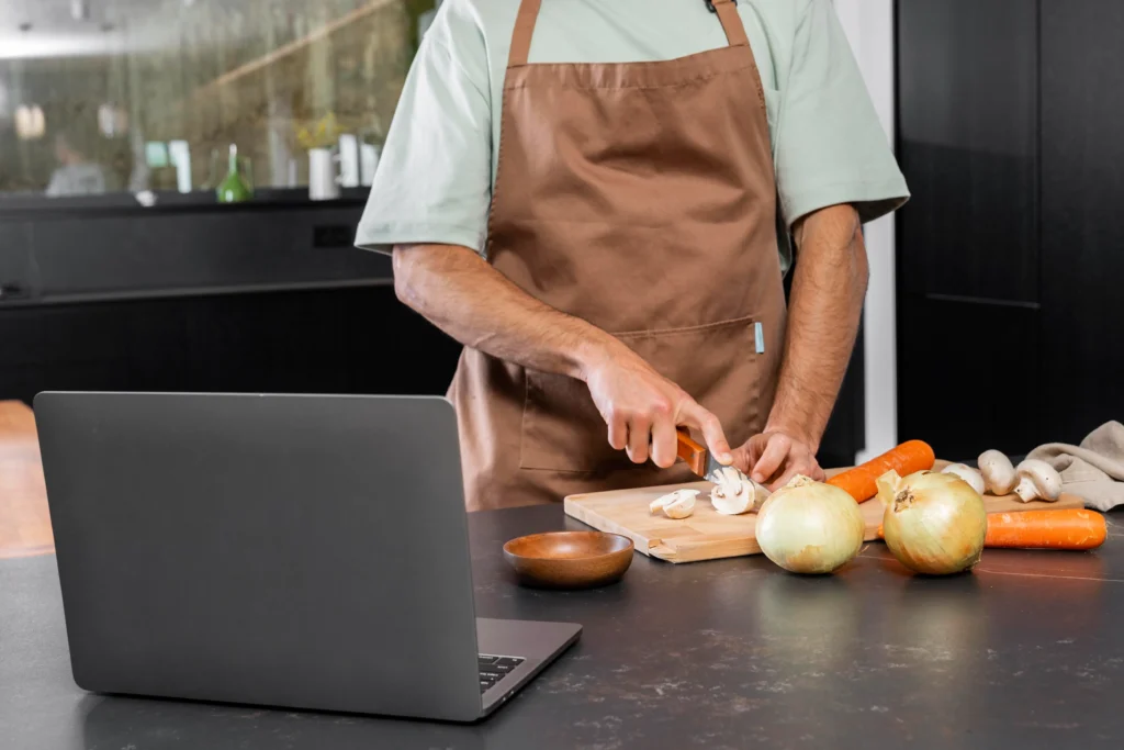A person wearing a brown apron chops fresh mushrooms on a wooden cutting board in a modern kitchen. An open laptop sits nearby on the dark countertop alongside whole onions and carrots.