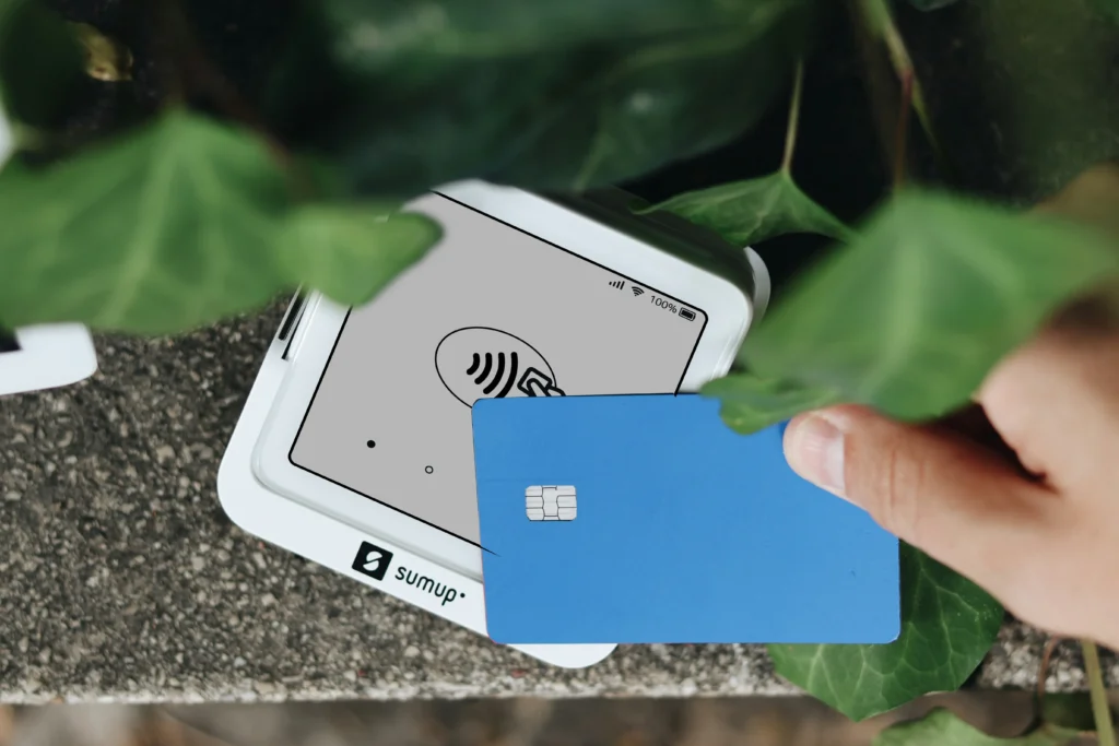 A hand holds a blue credit card above a white SumUp card reader to make a contactless payment. The portable reader displays a contactless signal icon and is surrounded by green plant leaves.
