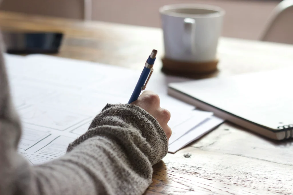 A person wearing a grey sweater holds a pen, working on paper laid out on a wooden table. A white coffee mug and a notebook rest nearby in the background.